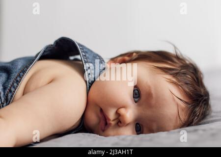 Portrait d'un bébé portant une barboteuse en denim couché sur une couverture grise à la maison. Bébé enfant aux yeux bleus fatigués, repose sur le lit dans la chambre, sur fond blanc Banque D'Images