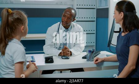 Medic discutant avec un petit enfant et un parent lors de la visite de rendez-vous au bureau. Spécialiste ayant une conversation avec la mère et la petite fille au sujet des services de soins de santé lors de la consultation. Banque D'Images