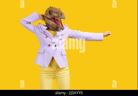 Femme en costume tendance et drôle masque de dinosaure dansant isolé sur fond jaune Banque D'Images