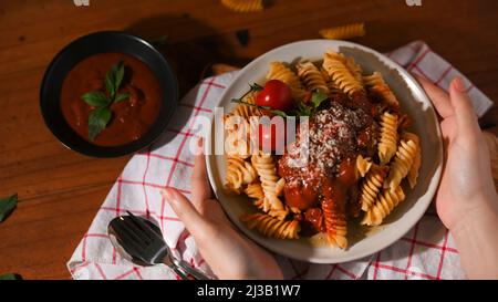 Vue de dessus, mains féminines servant une assiette de pâtes italiennes fusilli avec une sauce tomate spéciale sur une table à manger en bois. Banque D'Images