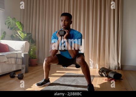 Jeune Afro-américain s'exerçant à la maison se concentrant tout en s'accroupidant avec un haltère dans le salon, l'entraînement à la maison Banque D'Images
