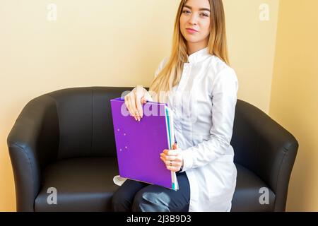 Portrait d'un jeune médecin caucasien avec des dossiers et des cheveux longs blonds portant un manteau blanc assis sur le canapé noir regardant l'appareil photo Banque D'Images
