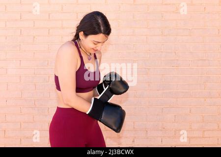 Une jeune femme caucasienne vêtue de leggings et d'un haut enfilant une paire de gants de boxe noirs. D'une main, elle ajuste le gant de l'autre Banque D'Images