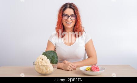 La femme caucasienne préfère une alimentation saine. REDHEAD fille choisit entre brocoli et beignes sur fond blanc. Banque D'Images