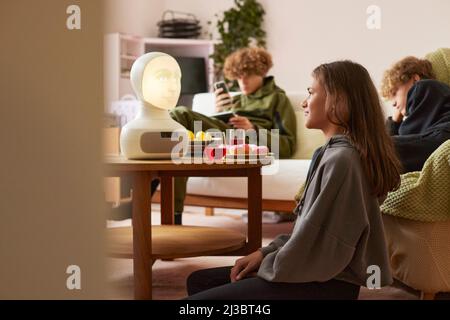 Frères et sœurs assis dans la salle de séjour et utilisant un assistant vocal robotique Banque D'Images