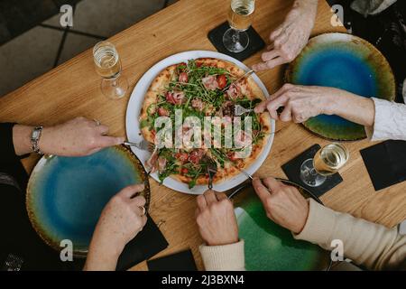 Vue de dessus de femmes âgées méconnues assis autour d'une table en bois avec des verres de champagne, creusant des fourchettes dans la pizza. Banque D'Images