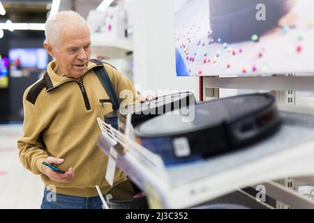 un homme âgé aux cheveux grisés qui regarde le robot hoover au comptoir dans la salle d'exposition du département d'hypermarché des appareils électriques Banque D'Images