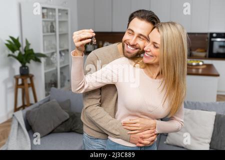 Un couple caucasien excité en amour se déplacer dans un nouvel appartement. Une femme joyeuse embrasse un gars barbu tenant les clés, le nouveau regard à la caméra et le sourire Banque D'Images