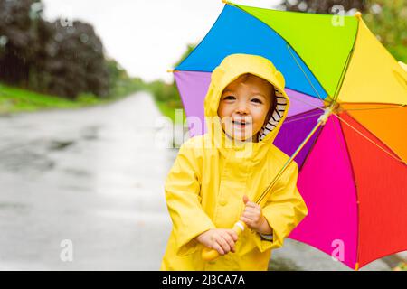 Drôle mignon bébé fille portant un manteau jaune imperméable et des bottes tenant un parapluie coloré jouant dans la pluie Banque D'Images