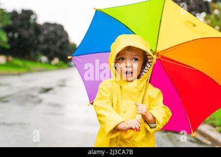 Drôle mignon bébé fille portant un manteau jaune imperméable et des bottes tenant un parapluie coloré jouant dans la pluie Banque D'Images