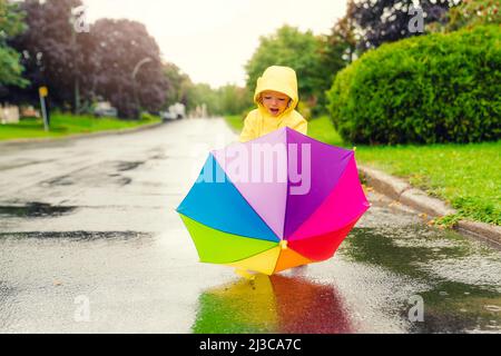 Drôle mignon bébé fille portant un manteau jaune imperméable et des bottes tenant un parapluie coloré jouant dans la pluie Banque D'Images