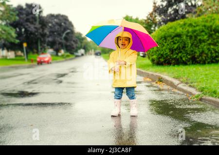 Drôle mignon bébé fille portant un manteau jaune imperméable et des bottes tenant un parapluie coloré jouant dans la pluie Banque D'Images