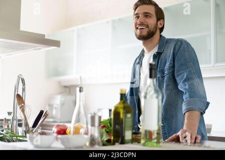 Beau jeune homme souriant se penchait sur le comptoir de la cuisine avec des légumes et regardant l'appareil photo. Portrait d'un homme décontracté heureux dans la cuisine avec des ingrédients e Banque D'Images