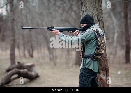 Le jeune homme pratique et vise à partir d'un pistolet à pompe élevé dans une armure de corps et de balaclava dans la forêt. Banque D'Images