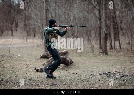 Le jeune homme pratique et vise à partir d'un pistolet à pompe élevé dans une armure de corps et de balaclava dans la forêt. Banque D'Images