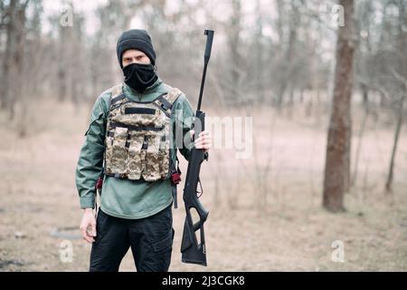 Le jeune homme se tient avec un pistolet à pompe dans sa main, élevé dans une armure de corps et de balaclava dans la forêt. Banque D'Images