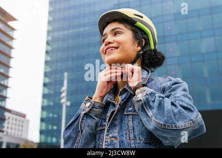 D'en bas jeune colombienne souriante dans une élégante tenue en denim attachant un casque de protection avant de monter à vélo dans la rue de la ville moderne Banque D'Images