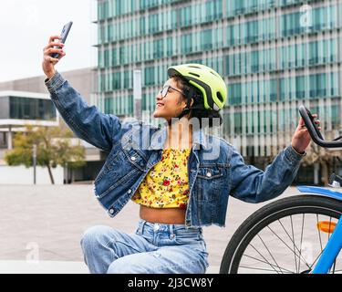Femme colombienne souriante en denim et casque de protection emportant un selfie sur un smartphone tout en prenant une pause à vélo dans la rue de la ville Banque D'Images