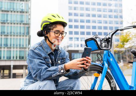 Femme colombienne souriante en denim et casque de protection parcourant un téléphone portable tout en prenant une pause à vélo dans la rue de la ville Banque D'Images