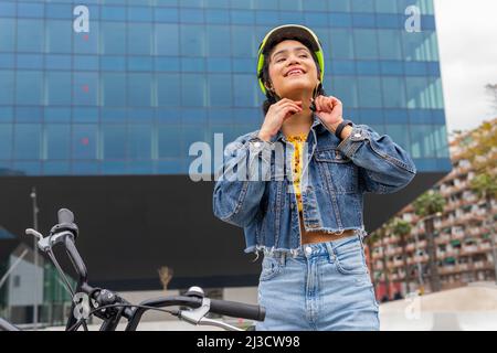 D'en bas jeune colombienne souriante dans une élégante tenue en denim attachant un casque de protection avant de monter à vélo dans la rue de la ville moderne Banque D'Images