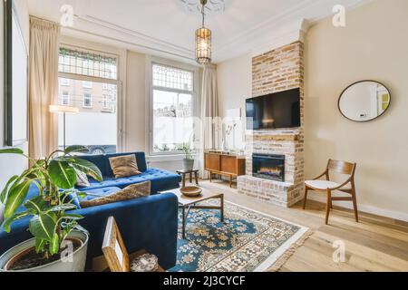 Confortable canapé bleu placé sur la moquette contre le mur avec TV et cheminée dans l'appartement élégant lumineux avec fenêtres et plantes en pot Banque D'Images