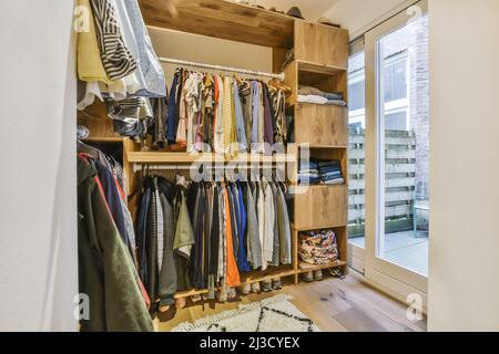 Garde-robe avec divers vêtements colorés et élégants accrochés sur des étagères avec des vêtements pliés près de la porte en verre dans l'appartement lumineux Banque D'Images