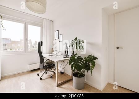 Chaise de bureau placée sur une table en bois avec écran d'ordinateur dans une chambre blanche spacieuse dans un plat conçu dans un style minimaliste Banque D'Images