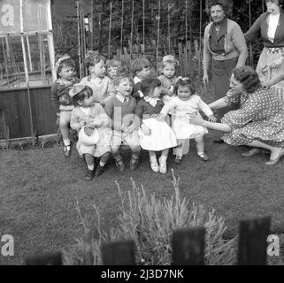 1961, hisotrical, une fête d'anniversaire de trois ans... à l'extérieur dans un jardin, une mère tente de faire s'asseoir sa petite fille impatiente avec les autres enfants pour une photo, Stockport, Manchester, Angleterre, Royaume-Uni. Banque D'Images
