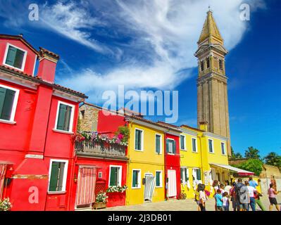 Venise (Burano), Italie - mai 9. 2019: Vue sur la rue sur une rangée de maisons jaune et rouge colorées, tour de l'église médiévale, ciel bleu, nuages moelleux Banque D'Images
