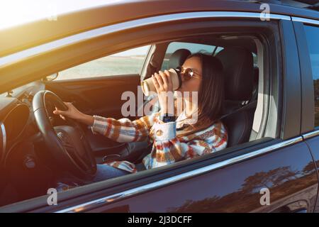 Jeune belle femme élégante conduisant sa nouvelle voiture et buvant un café par temps ensoleillé. Concept de location de voiture Banque D'Images