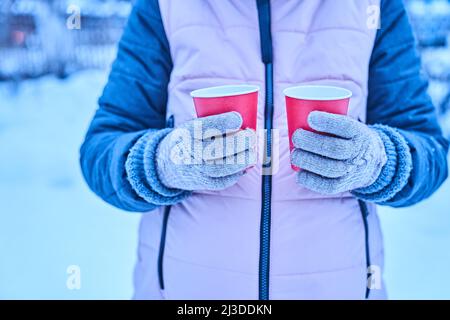 Une femme en gants tient dans ses mains deux tasses en papier rouge de boisson chaude en hiver sur la nature, un thé chaud en saison froide, une promenade et un petit voyage en hiver. Photo de haute qualité Banque D'Images