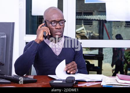 un bel homme dans un polo bleu assis au bureau parlant sur le téléphone portable tout en vérifiant un document. Banque D'Images
