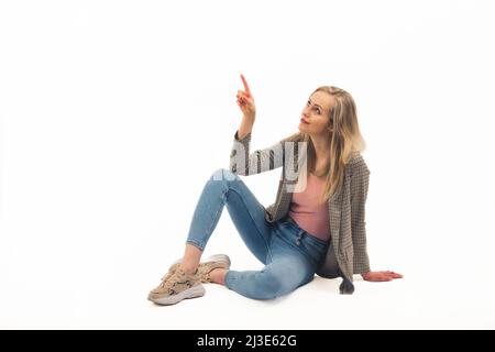 Agréable blanche blonde fille assise sur le sol et avec un doigt pointant vers le haut plein plan studio tourné blanc arrière-plan copie espace. Photo de haute qualité Banque D'Images