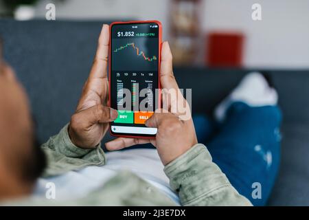 Homme multiracial allongé sur un canapé et tenant un téléphone avec l'application de bourse Banque D'Images