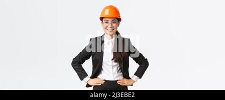 Femme asiatique sourit, ingénieur de construction, architecte en costume noir et casque de sécurité, qui se tient satisfaite de son travail Banque D'Images