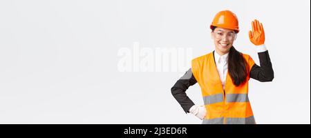 Sympathique femme asiatique souriante ingénieur de construction, architecte dans casque de sécurité, gants et lunettes de main, en disant bonjour, saluant les investisseurs ou Banque D'Images