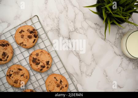 Vue de dessus, plat, petits gâteaux aux pépites de chocolat fraîchement cuits sur une grille de refroidissement avec un verre de lait, une plante de décoration et un espace de copie sur fond de marbre blanc Banque D'Images