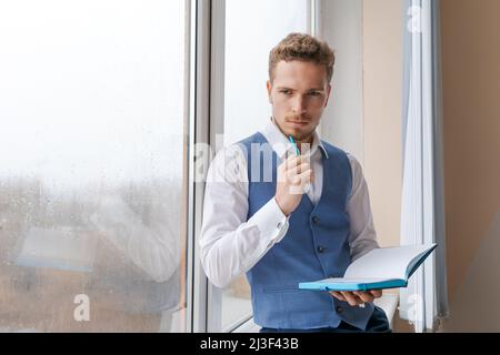 Un jeune homme d'affaires barbu travaille au bureau, prend des notes dans un carnet, analyse les informations, prépare un plan d'affaires tout en étant assis près d'une fenêtre sur le rebord de la fenêtre. Entrepreneur indépendant travaillant à domicile. Banque D'Images