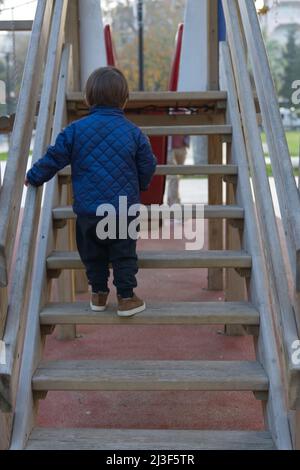 Petit garçon caucasien vue arrière dans le parc sur le terrain de jeu dans la glissade rouge blanc portant un manteau d'hiver en automne. Banque D'Images
