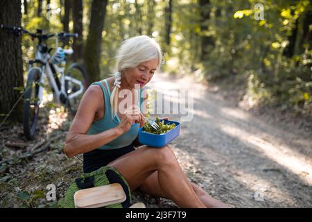 Heureuse femme biker senior se reposer, s'asseoir et manger des encas à l'extérieur dans la forêt. Banque D'Images