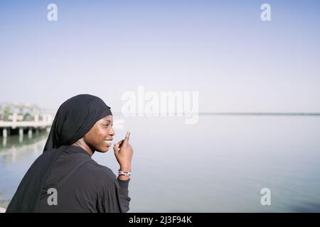 Une jeune fille musulmane africaine souriante, assise détendue au bord de la mer et pointant vers l'horizon, symbolise la confiance et l'espoir dans l'avenir des jeunes générations Banque D'Images