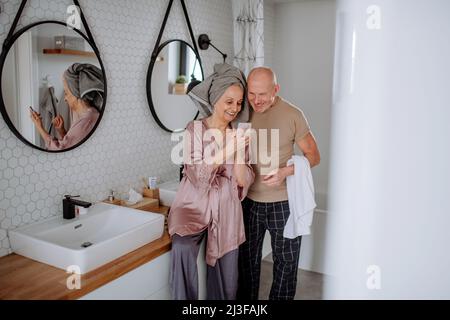 Couple senior amoureux dans la salle de bains, utilisant un smartphone, concept de routine du matin. Banque D'Images