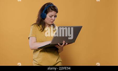 Femme décontractée portant un casque sans fil tenant un ordinateur portable et tapant sur un clavier regardant l'écran debout en studio. Personne détendue écoutant de la musique sur des écouteurs à l'aide d'un ordinateur portable. Banque D'Images