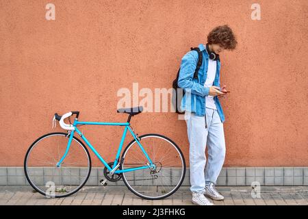 Jeune élève aux cheveux bouclés avec un casque posé sur un mur à l'extérieur, utilisant un téléphone cellulaire à côté de son vélo bleu Banque D'Images