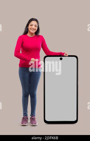 Jeune femme souriante se penchée sur un grand smartphone et pointant Banque D'Images