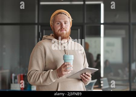 Portrait du contenu moderne barbu rouge homme dans un chapeau debout avec une tablette et thermos Cup dans le bureau Banque D'Images