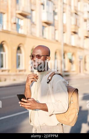 Un jeune homme bald afro-américain pensif qui écoute de la musique dans des écouteurs tout en utilisant des gadgets dans la rue Banque D'Images