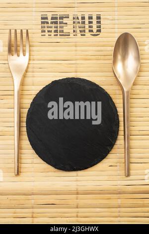 Maquette de couverture de menu avec un tapis de table en bambou, des couverts peints en or et un carrelage en pierre noire Banque D'Images