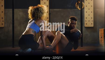 Construire un noyau plus fort à chaque torsion. Photo d'un jeune homme faisant des torsades russes avec une balle lestée et l'aide d'un entraîneur dans une salle de gym. Banque D'Images