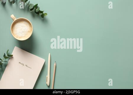 Table de bureau moderne avec carnet en papier, tasse de café, stylo, crayon et feuilles d'eucalyptus sur fond vert. Style minimaliste Banque D'Images